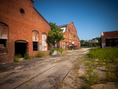 Abandoned brick buildings with broken windows and overgrown vegetation under a clear blue sky.