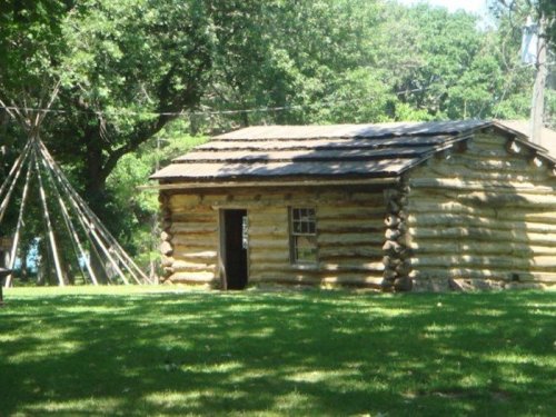 A rustic log cabin surrounded by trees, with a teepee structure nearby on a grassy area.