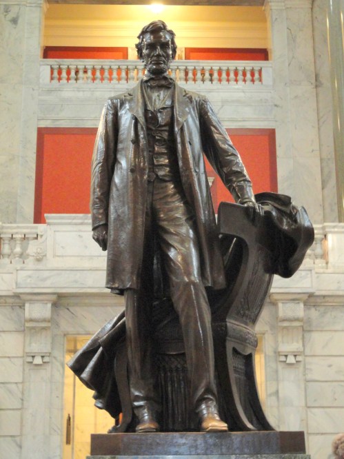 A bronze statue of a man in a suit, standing confidently with one hand on a chair, set in a grand interior.
