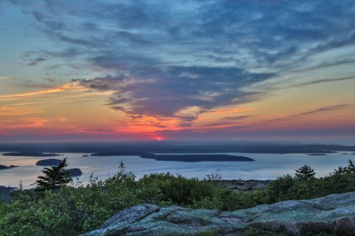 A vibrant sunset over a calm bay, with islands silhouetted against a colorful sky and lush greenery in the foreground.