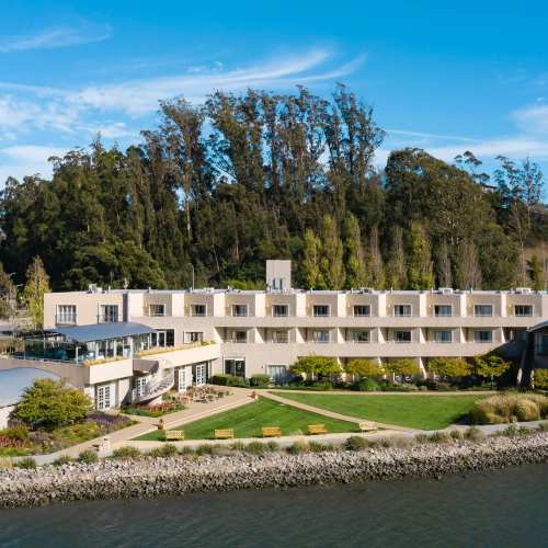 A waterfront hotel with a modern design, surrounded by greenery and trees, under a clear blue sky.