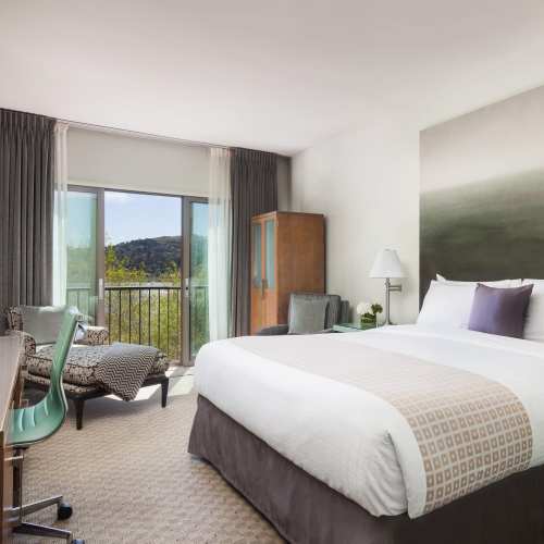 A modern hotel room featuring a king bed, seating area, desk, and large windows with a view of greenery outside.