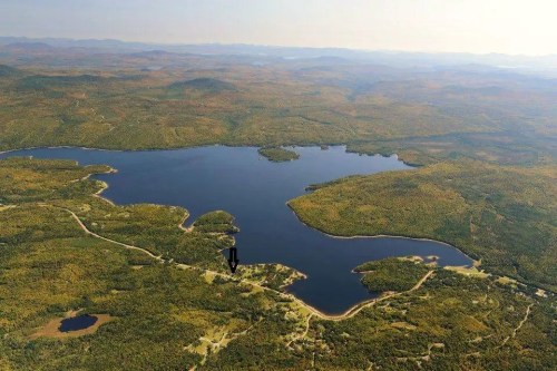 Aerial view of a large lake surrounded by dense forests and rolling hills under a clear sky.