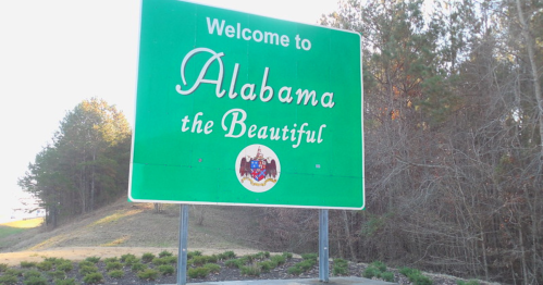 Welcome sign for Alabama, featuring the phrase "Alabama the Beautiful" and the state seal. Green background with trees.