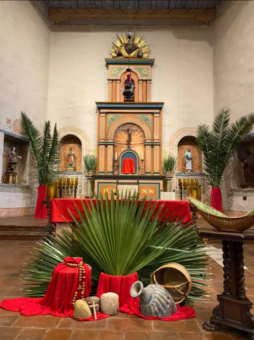 A decorated altar with palm leaves, red fabric, and religious statues in a serene interior setting.