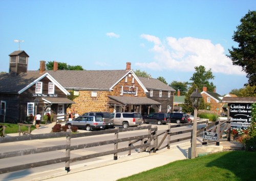 A quaint shopping area featuring stone buildings, parked cars, and a wooden fence under a clear blue sky.
