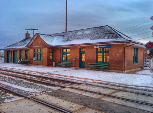 A brick train station with green benches, surrounded by snow and railway tracks under a cloudy sky.