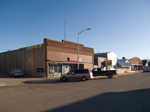 A street view of a small town with brick buildings, a clear sky, and parked cars along the road.