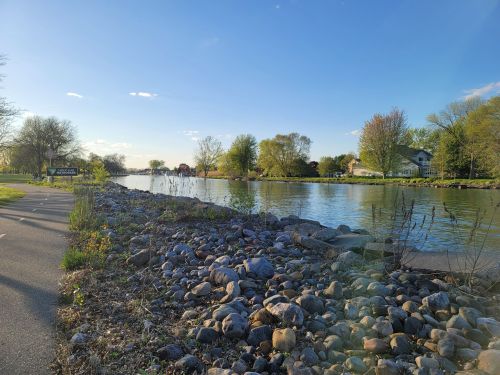 A serene river scene with a rocky shore, trees, and a clear blue sky, alongside a walking path.