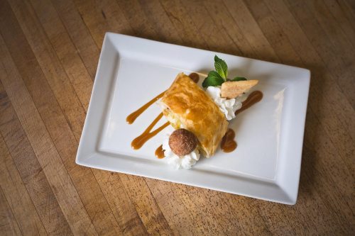 A slice of pastry dessert topped with whipped cream, caramel drizzle, a mint leaf, and a cookie on a white plate.