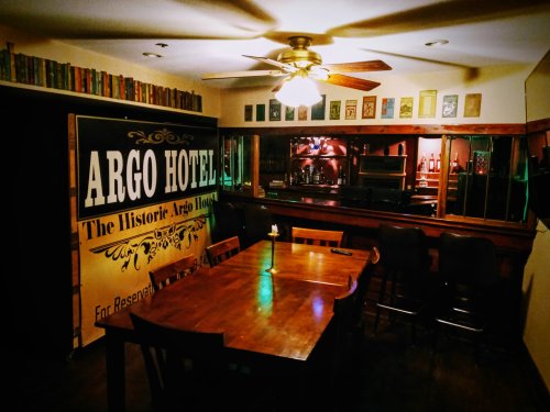 Interior of a dimly lit room featuring a large wooden table, bar area, and a banner for the Argo Hotel on the wall.
