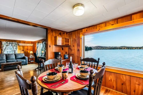 Cozy dining area with a table set for four, overlooking a serene lake through a large window. Warm wooden interior.