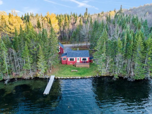 Aerial view of a red cabin by a lake, surrounded by lush green trees and autumn foliage.