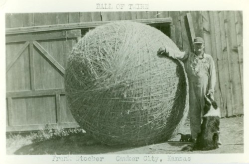 A man stands next to a giant ball of twine with a dog at his feet, in front of a wooden barn in Kansas.