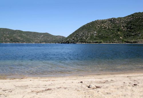 A serene beach scene with clear water, sandy shore, and green hills in the background under a clear blue sky.