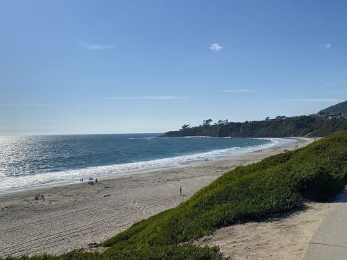 A serene beach scene with gentle waves, sandy shore, and green hills under a clear blue sky.