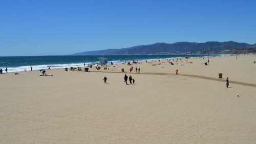 A sunny beach scene with people walking on the sand, waves crashing, and mountains in the background.