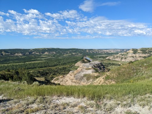 A scenic landscape featuring rolling hills, green valleys, and a blue sky with scattered clouds.