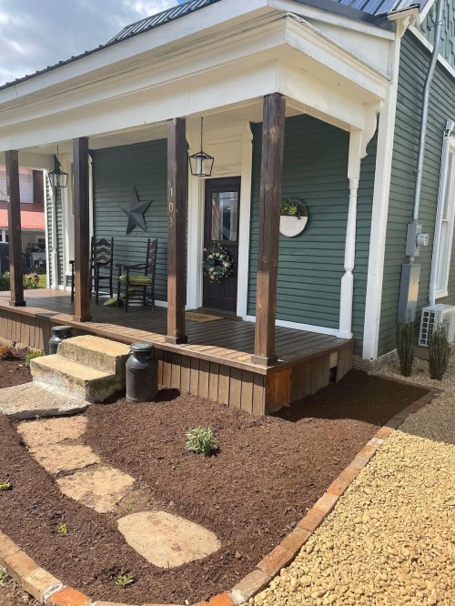 A charming green house with a porch, wooden pillars, and a landscaped yard featuring mulch and stone pathways.