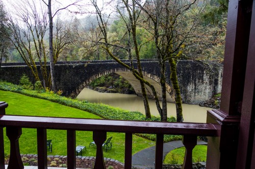 A stone bridge arches over a muddy river, surrounded by lush greenery and trees, viewed from a balcony.