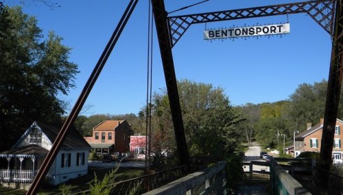 View of a historic bridge with a sign reading "Bentonport," surrounded by trees and quaint buildings under a clear blue sky.