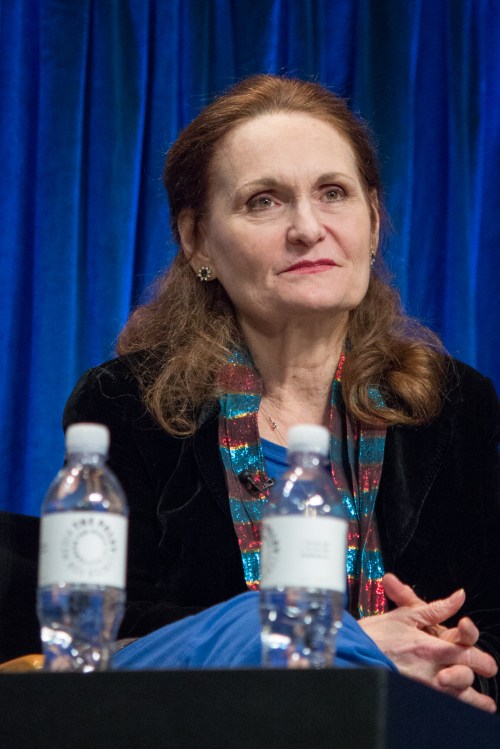 A woman with long brown hair, wearing a black jacket and colorful scarf, sits thoughtfully at a panel discussion.