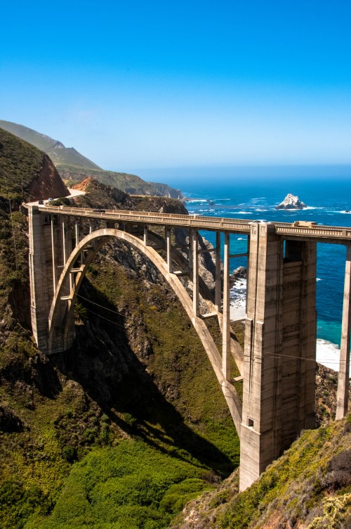 A scenic view of a large concrete bridge spanning a coastal canyon, with ocean waves and mountains in the background.