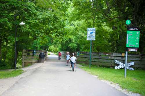 Children riding scooters on a paved path surrounded by greenery, with signs for a trail and a railroad crossing.