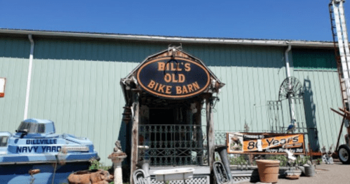 Sign for "Bill's Old Bike Barn" with vintage bikes and decor outside a green building under a clear blue sky.