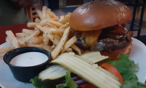 A juicy burger with bacon and toppings, served with crispy fries, pickles, lettuce, and tomato on a white plate.