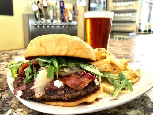 A juicy burger topped with bacon, onions, and arugula, served with fries and a pint of beer in a bar setting.
