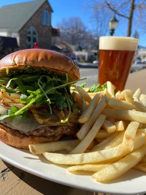 A gourmet burger topped with greens and onions, served with fries and a glass of amber beer on a sunny day.