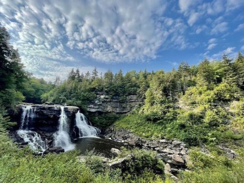 A scenic waterfall surrounded by lush greenery and a blue sky with clouds.