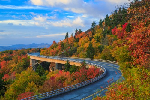 A winding road curves through vibrant autumn foliage under a blue sky with scattered clouds.