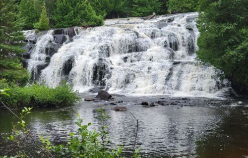 A serene waterfall cascades over rocks, surrounded by lush greenery and reflecting in a calm pool below.