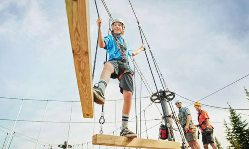 A child balances on a wooden beam in an outdoor ropes course, with two adults observing in the background.