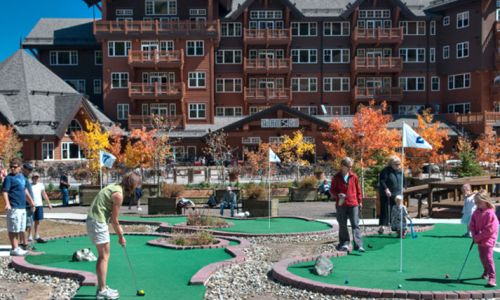 A mini-golf course with players of various ages, surrounded by colorful autumn trees and a large lodge in the background.