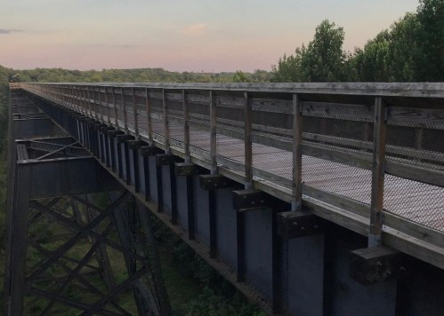 A wooden bridge spans a lush green landscape, with trees lining the horizon under a pastel sky.