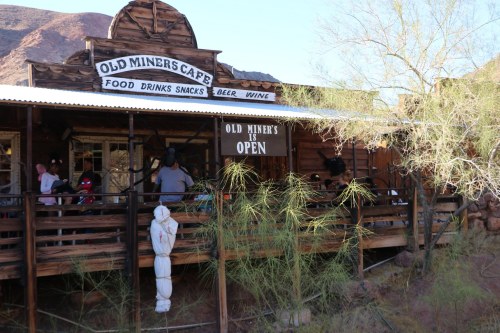 A rustic café with a wooden exterior, featuring a sign that says "Old Miner's is Open" and people on the porch.