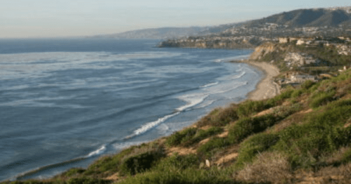A scenic coastal view with gentle waves, sandy beach, and lush greenery along the shoreline under a clear sky.