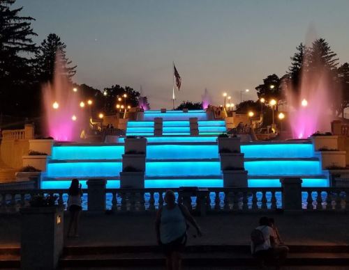 A brightly lit fountain with blue and pink lights, cascading steps, and an American flag at dusk.