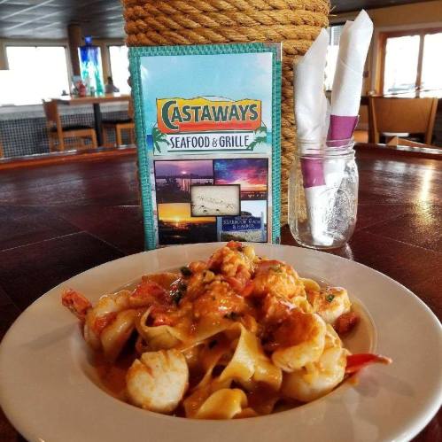 A plate of seafood pasta with shrimp and scallops, served at Castaways Seafood & Grille, with a menu in the background.