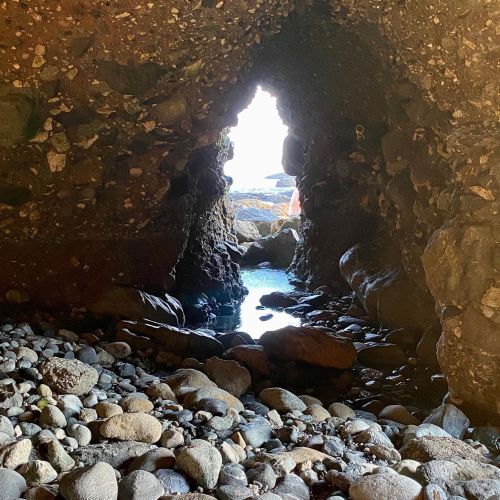 A rocky cave opening revealing a bright light and water beyond, with smooth pebbles scattered on the ground.