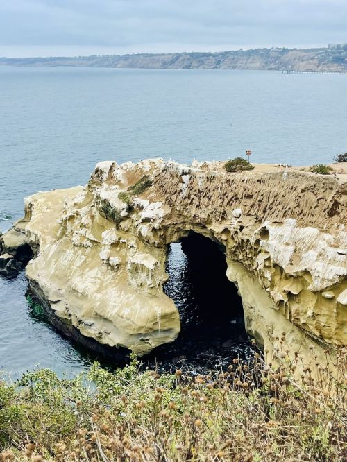 A coastal rock formation with an arch, overlooking calm waters and distant cliffs under a cloudy sky.