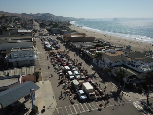 Aerial view of a beachside street market with vendors, cars, and a scenic ocean backdrop on a sunny day.