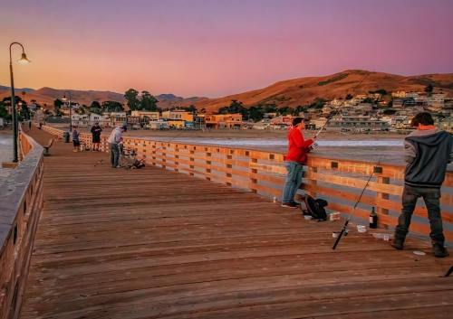 Fishermen on a wooden pier at sunset, with hills and houses in the background under a colorful sky.