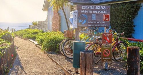 Sign for coastal access with bicycles parked nearby, surrounded by greenery and a pathway leading to the beach.