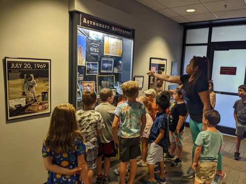 A group of children gathers around a display case, while an adult points out items related to astronauts.