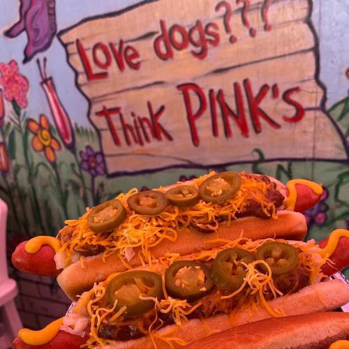 Two hot dogs topped with cheese, jalapeños, and onions, in front of a colorful mural with the text "Love dogs??? Think PINK's."