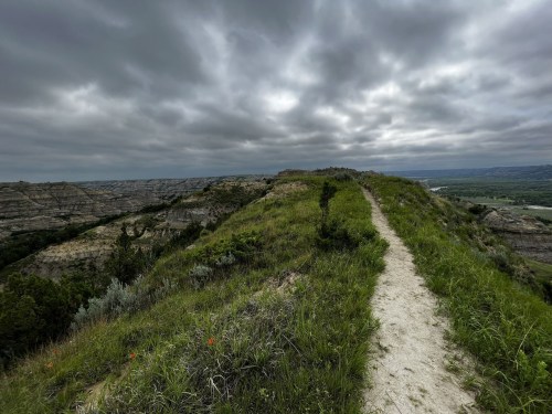 A winding path leads through lush green grass atop a rocky ridge under a cloudy sky.
